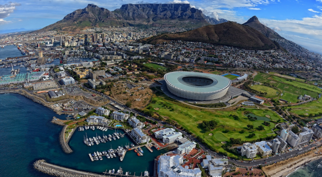 Aerial view of Cape Town featuring the Cape Town Stadium, city buildings, Table Mountain in the background, and a marina with boats in the foreground under a partly cloudy sky.
