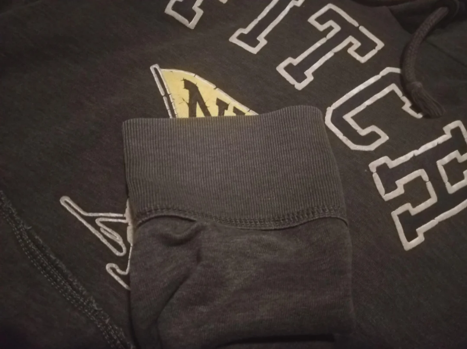 A close-up of a dark grey sweatshirt with white outlined letters and a folded sleeve. Part of a yellow and black graphic is visible beneath the lettering.