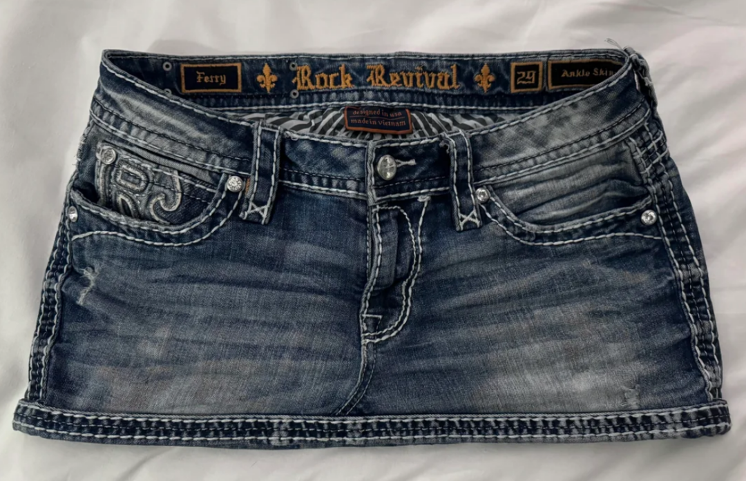 A short, distressed blue denim mini skirt with white stitching, belt loops, and front pockets, featuring "Rock Revival" branding on the waistband.