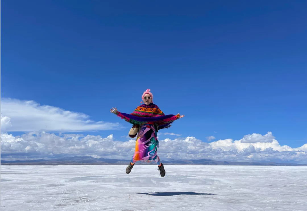 A person in colorful clothing and sunglasses jumps mid-air on a vast, white salt flat under a bright blue sky with scattered clouds and distant mountains.