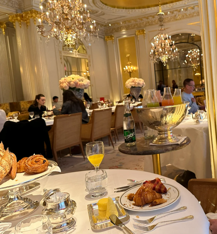 Elegant restaurant with ornate chandeliers, gold accents, and floral centerpieces. People are dining at round tables set with pastries, drinks, and cutlery. A plate of food and a glass of juice are in focus in the foreground.