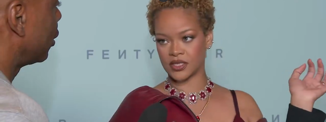 A woman with short curly hair, wearing a burgundy dress and a jeweled necklace, speaks at a Fenty event. She stands in front of a blue backdrop with "Fenty" text, holding one hand up as a microphone is held toward her.