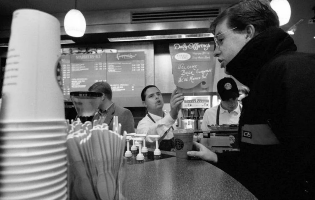 A customer orders at a busy coffee shop counter while staff prepare drinks, with stacks of cups, stirrers, and a menu board visible in the background; the scene is in black and white.