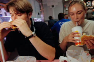 A young man and woman sit at a fast-food restaurant table, eating burgers and drinking from cups. Both appear relaxed and focused on their food, surrounded by other diners in the background.