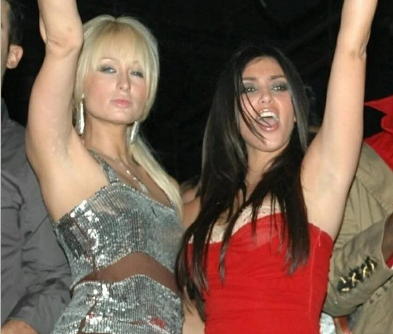 Two women pose together at a party, both raising their arms. One wears a silver sequin dress and has blonde hair with bangs; the other wears a red strapless dress and has long dark hair. Both appear to be celebrating or dancing.