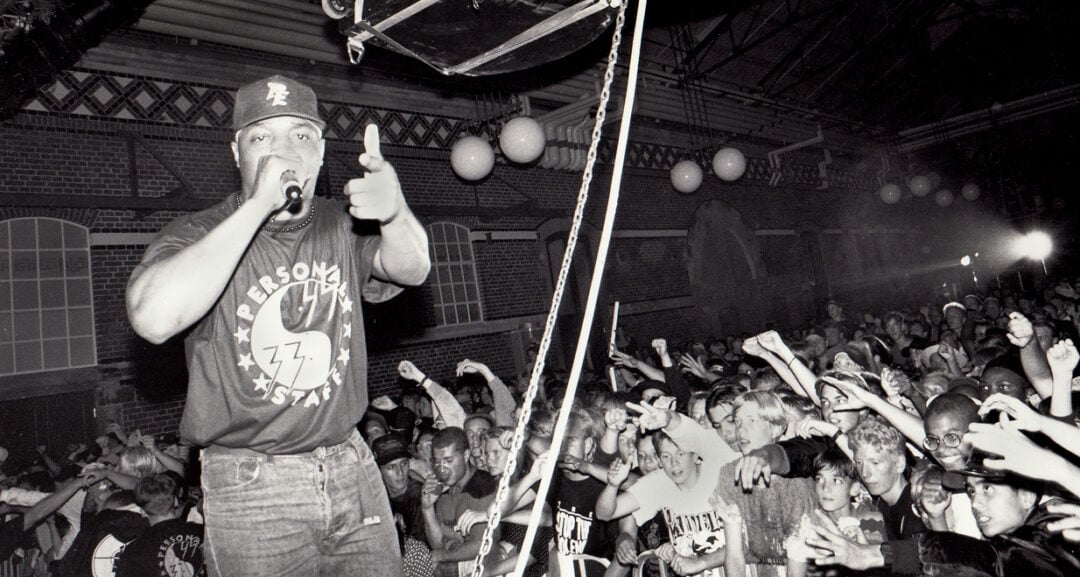 A rapper performs energetically on stage, holding a microphone and gesturing to a cheering, packed crowd in a large indoor venue with exposed brick walls and round hanging lights.