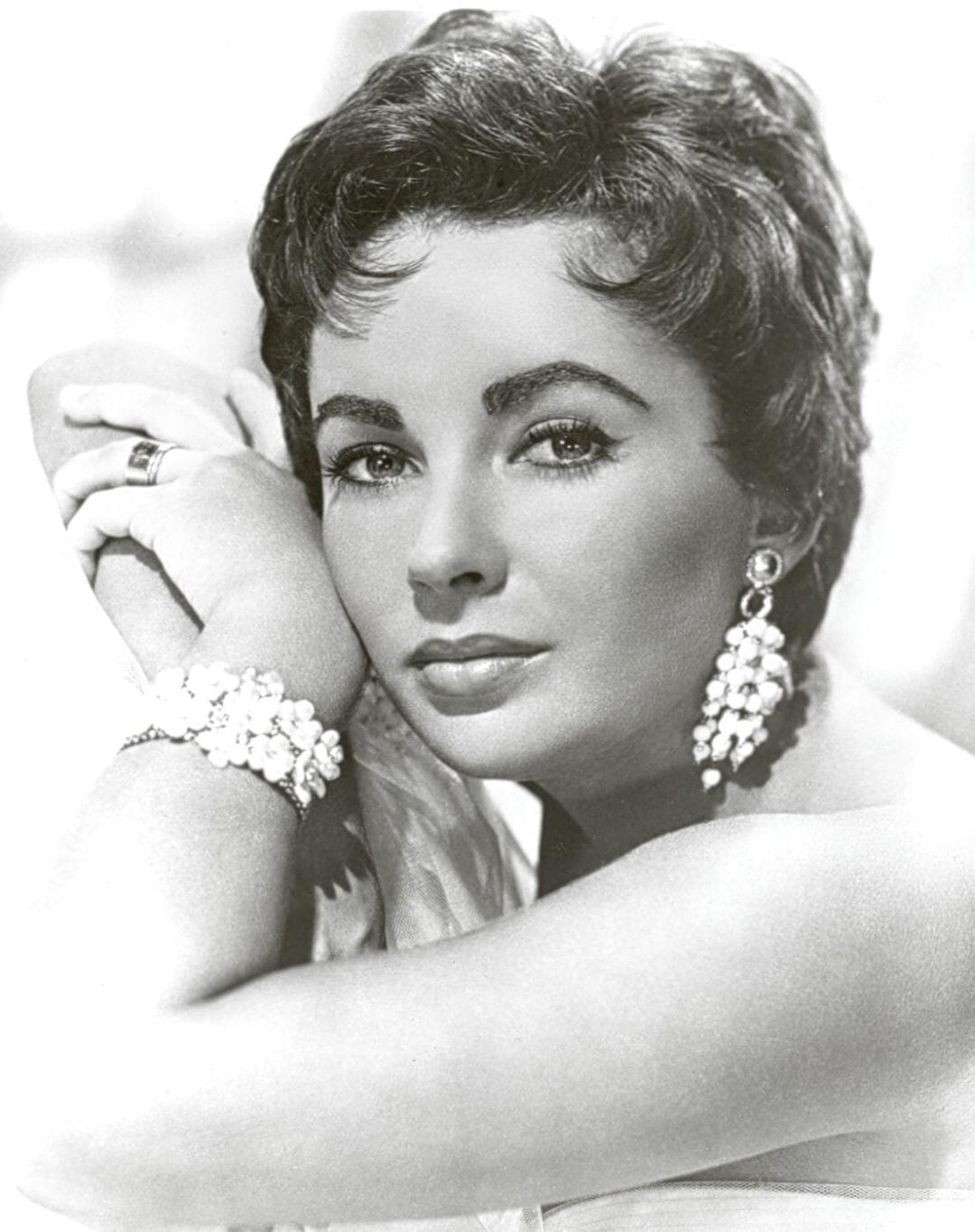 A glamorous woman with short, dark hair poses with her head resting on her arm. She wears sparkling earrings, a matching beaded bracelet, and a ring, looking directly at the camera with a soft, confident expression.
