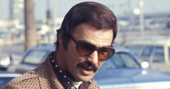 A man with short dark hair, a mustache, and sideburns wears large, brown sunglasses and a brown patterned jacket over a dark shirt. He is outdoors with cars and a palm tree in the blurred background.