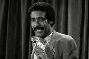 A man with a mustache and curly hair stands in front of dark curtains, holding a microphone and speaking, dressed in a suit jacket and collared shirt. The image is in black and white.