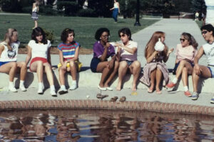 A group of eight young people sit on the edge of a brick fountain, talking and laughing on a sunny day, with shoes off and feet near the water. Two more people walk in the background near trees and a lawn.