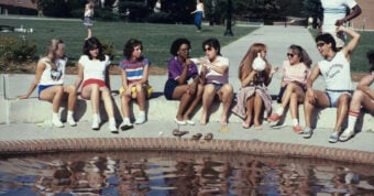 A group of eight young people sit on the edge of a brick fountain, talking and laughing on a sunny day, with shoes off and feet near the water. Two more people walk in the background near trees and a lawn.