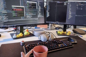 A workspace with two large monitors displaying code, a mechanical keyboard, headphones, a coffee mug, and several rubber ducks on the desk. The setup appears organized and tech-focused.