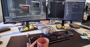 A workspace with two large monitors displaying code, a mechanical keyboard, headphones, a coffee mug, and several rubber ducks on the desk. The setup appears organized and tech-focused.