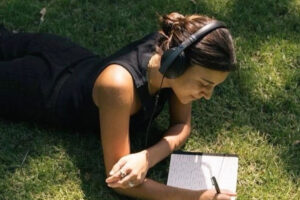 A person lies on the grass wearing headphones, writing in a notebook with a pen. The person is dressed in black and appears focused, enjoying a sunny day outdoors.