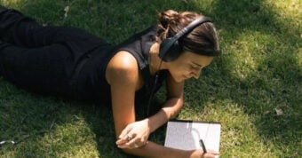 A person lies on the grass wearing headphones, writing in a notebook with a pen. The person is dressed in black and appears focused, enjoying a sunny day outdoors.