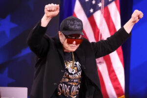A person wearing sunglasses, a black hat, and a dark jacket raises both arms with clenched fists on stage. An American flag is visible in the background.
