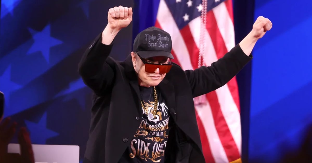 A person wearing sunglasses, a black hat, and a dark jacket raises both arms with clenched fists on stage. An American flag is visible in the background.