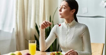 A woman in a white zip-up top sits at a table, holding a fork with food, and looks thoughtfully out the window. There is a plate of food and a glass of orange juice in front of her.