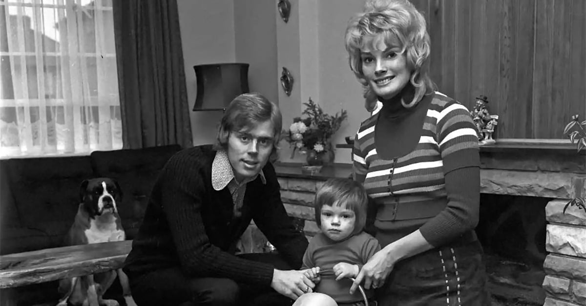 A man and woman pose with a young child in a living room. The man kneels, holding the child’s hand, while the woman stands beside them, smiling. A dog sits on a couch in the background. The room has a fireplace and curtains.