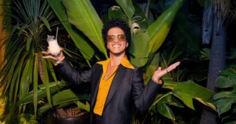 A person wearing sunglasses, a yellow shirt, and a dark suit smiles and holds a cocktail while posing in front of large tropical plants with arms spread wide.