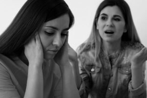 Two women sit together; one looks upset with her head in her hands, while the other gestures and appears to be speaking emotionally. The mood is tense. The image is in black and white.
