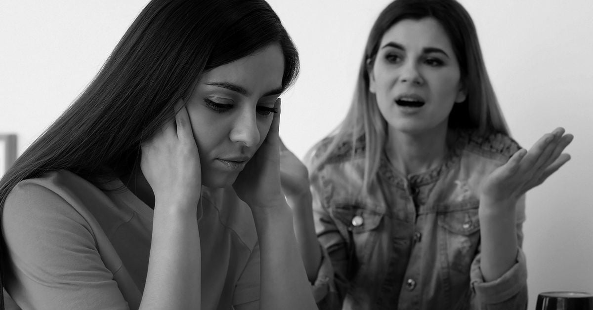Two women sit together; one looks upset with her head in her hands, while the other gestures and appears to be speaking emotionally. The mood is tense. The image is in black and white.