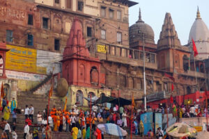 A large crowd gathers on the steps of a ghat along the Ganges River in Varanasi, India, with historic temples and buildings in the background. People wear colorful clothing and some carry orange flags.
