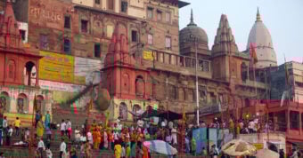 A large crowd gathers on the steps of a ghat along the Ganges River in Varanasi, India, with historic temples and buildings in the background. People wear colorful clothing and some carry orange flags.