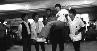 A black-and-white photo of boxer Muhammad Ali playfully holding up a smiling member of The Beatles, while the other three band members stand around them, all laughing and looking at each other.