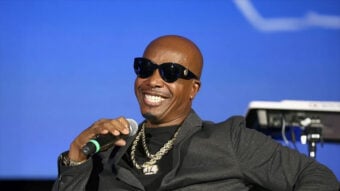 A man wearing dark sunglasses, a black jacket, and gold chains is smiling and holding a microphone, seated against a blue background with musical equipment visible behind him.