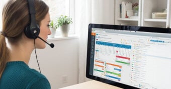A woman wearing a headset sits at a desk and looks at a computer screen displaying a colorful calendar and scheduling software in a bright home office.