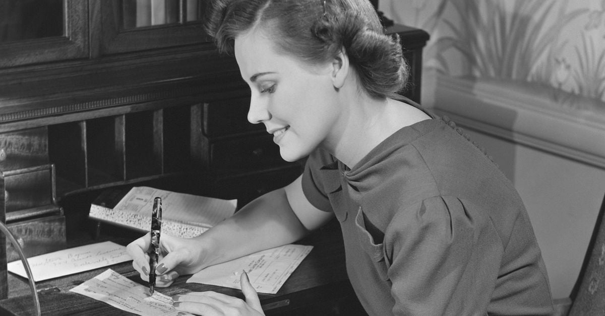 A woman in vintage clothing sits at a wooden desk, smiling as she writes with a fountain pen on paper. There are letters and an open notebook on the desk, and the scene is in black and white.