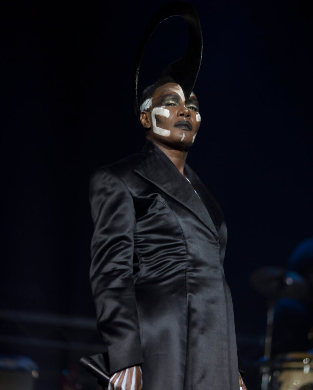 A person in a dramatic black coat stands on stage, wearing bold white and black face paint and a large circular headpiece, with their hand painted in white stripes, against a dark background.