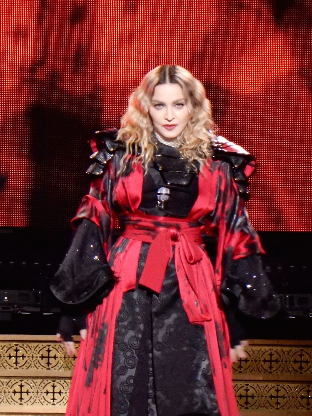 A woman with blonde, wavy hair stands on stage, wearing a black and red elaborate robe with wide sleeves and a sash, in front of a bright red and black background.