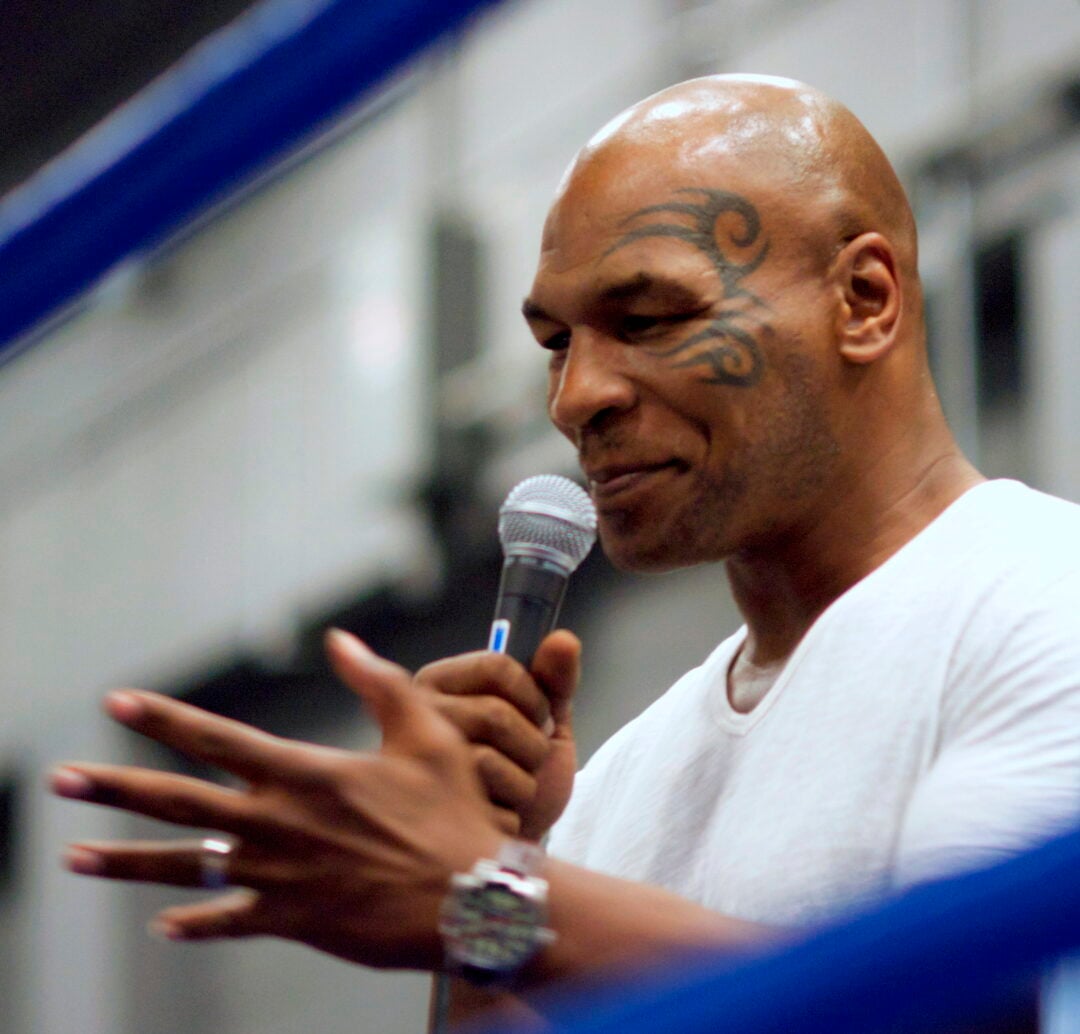 A man with a tribal face tattoo and a shaved head speaks into a microphone while gesturing with his hand, wearing a white shirt and a watch, with blue lines blurred in the foreground.