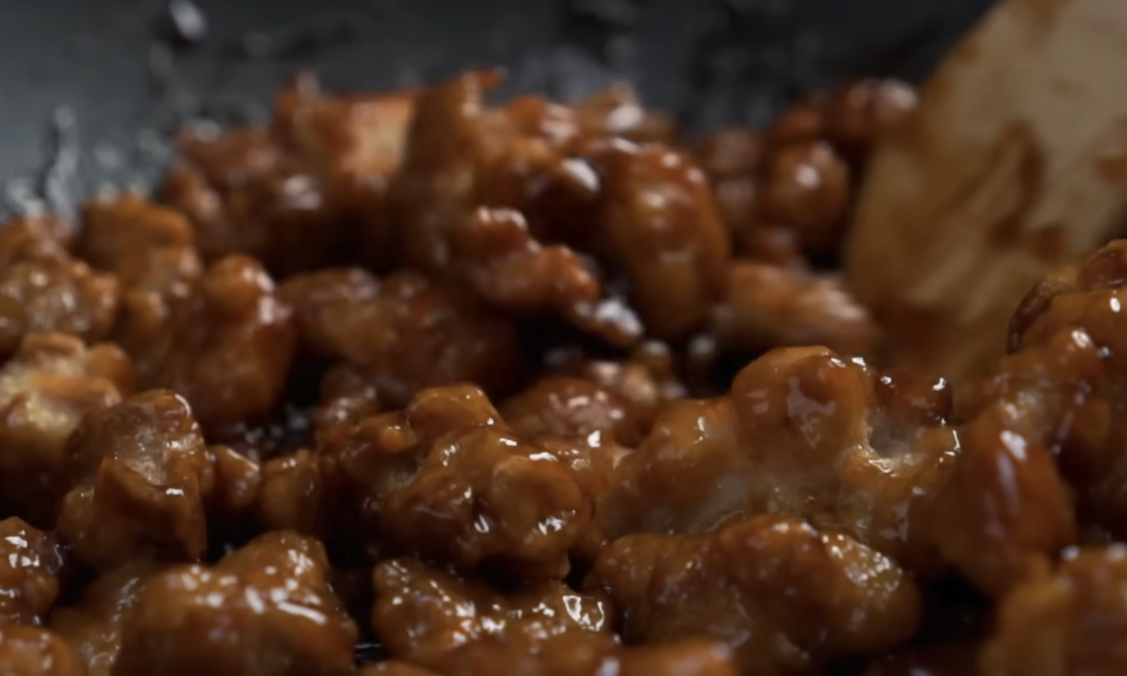 Bite-sized pieces of fried chicken coated in a thick, dark glaze being stirred in a pan with a wooden spoon.