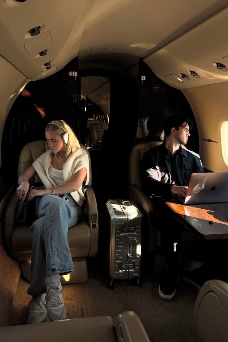A woman with headphones sits by the aisle, looking down, while a man works on a laptop by the window in a private jet. A silver suitcase is placed between them. Warm lighting fills the cabin.