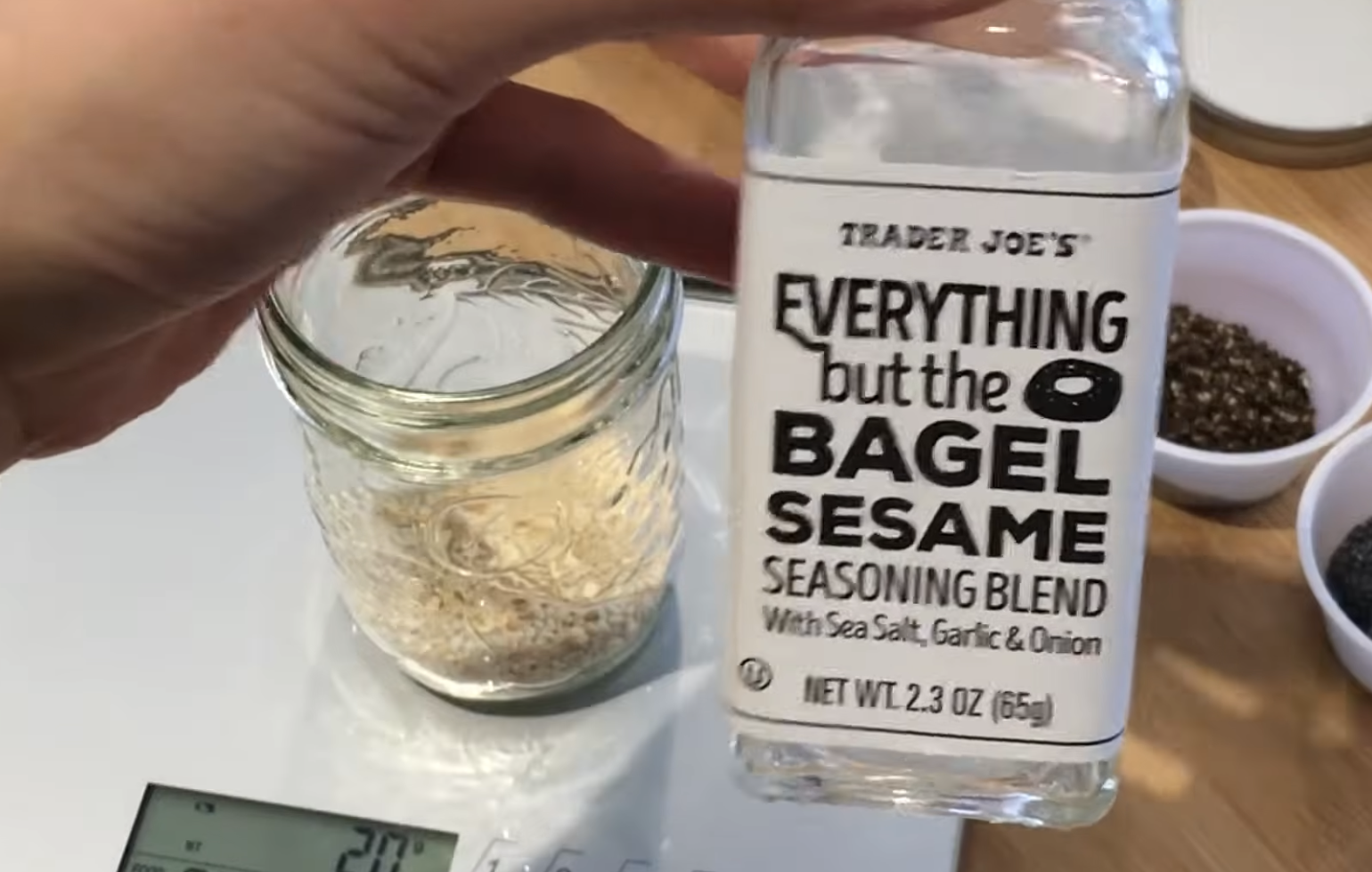 A hand holds a bottle of Trader Joe’s Everything but the Bagel Sesame Seasoning Blend next to a glass jar on a kitchen scale.