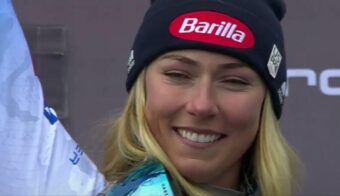 A woman with long blonde hair, wearing a black beanie with a red "Barilla" logo, smiles while holding up a flag or banner. She is dressed in winter clothing and appears to be at an event.