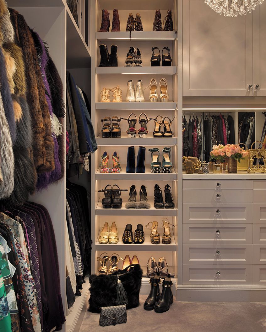 A luxurious walk-in closet with shelves displaying high-heeled shoes, hanging fur coats and clothes, white cabinets with drawers, a chandelier, and a countertop with flowers and decorative items.