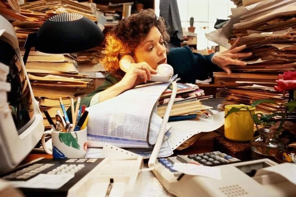 A woman surrounded by towering stacks of papers talks on the phone while searching through files at a cluttered desk filled with office supplies, a computer, and a calculator.
