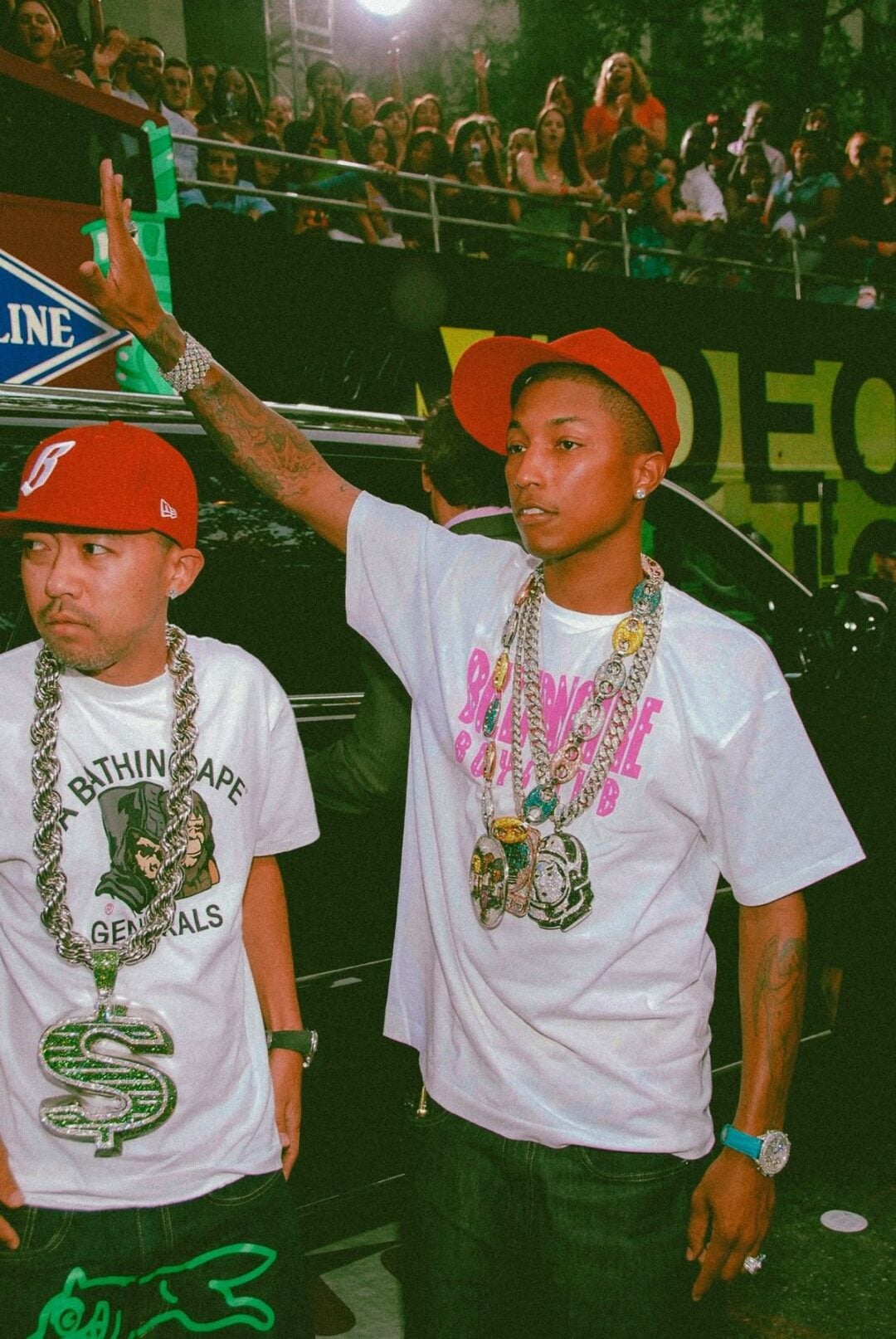 Two men stand outdoors at an event. Both wear red caps, white graphic t-shirts, and large chains. The man on the right raises his hand, while the crowd in the background watches. Bright lights illuminate the scene.
