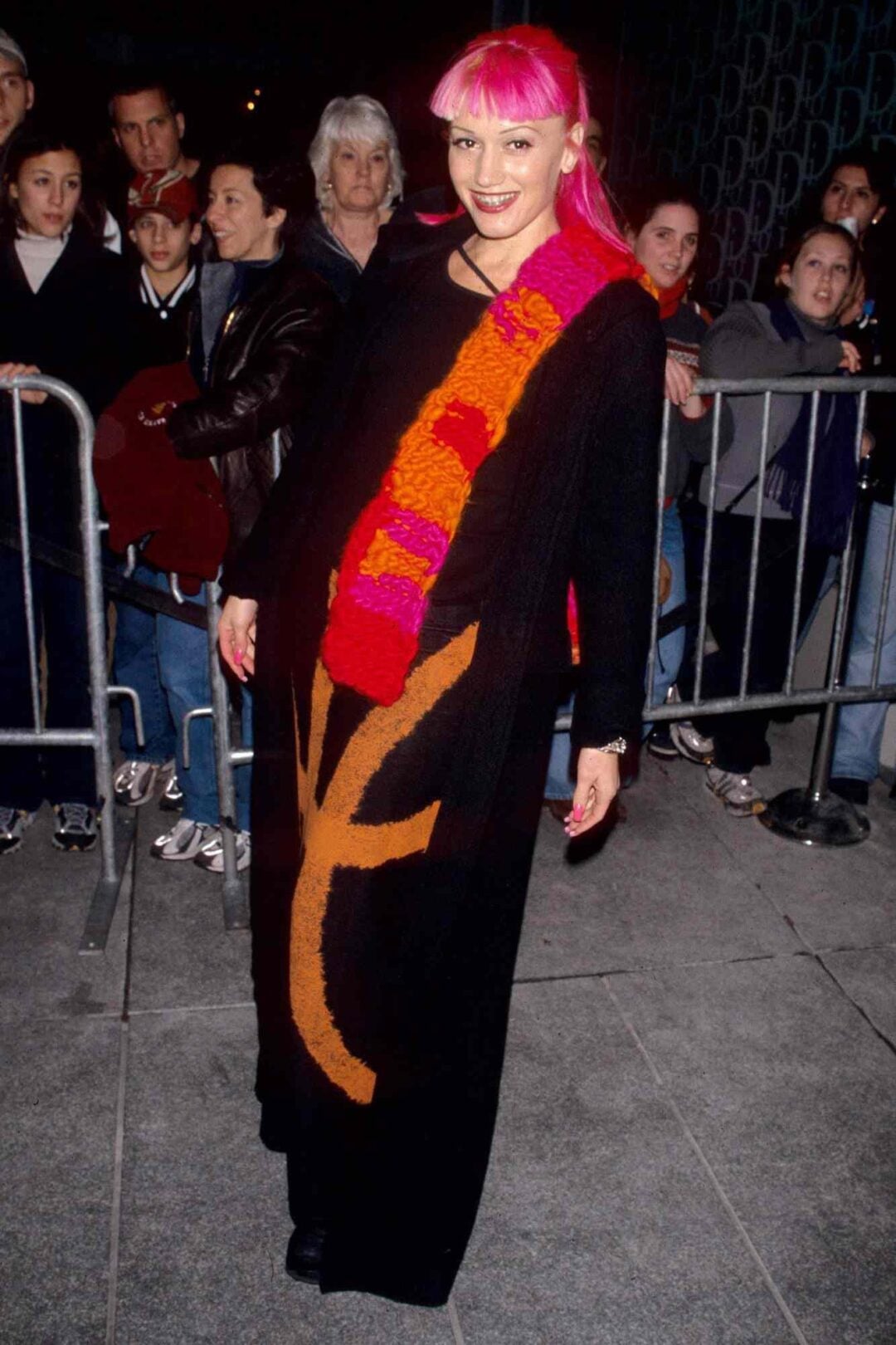 A woman with bright pink hair smiles while wearing a long black dress with bold orange and red patterns, standing in front of a crowd behind metal barriers on a city sidewalk at night.
