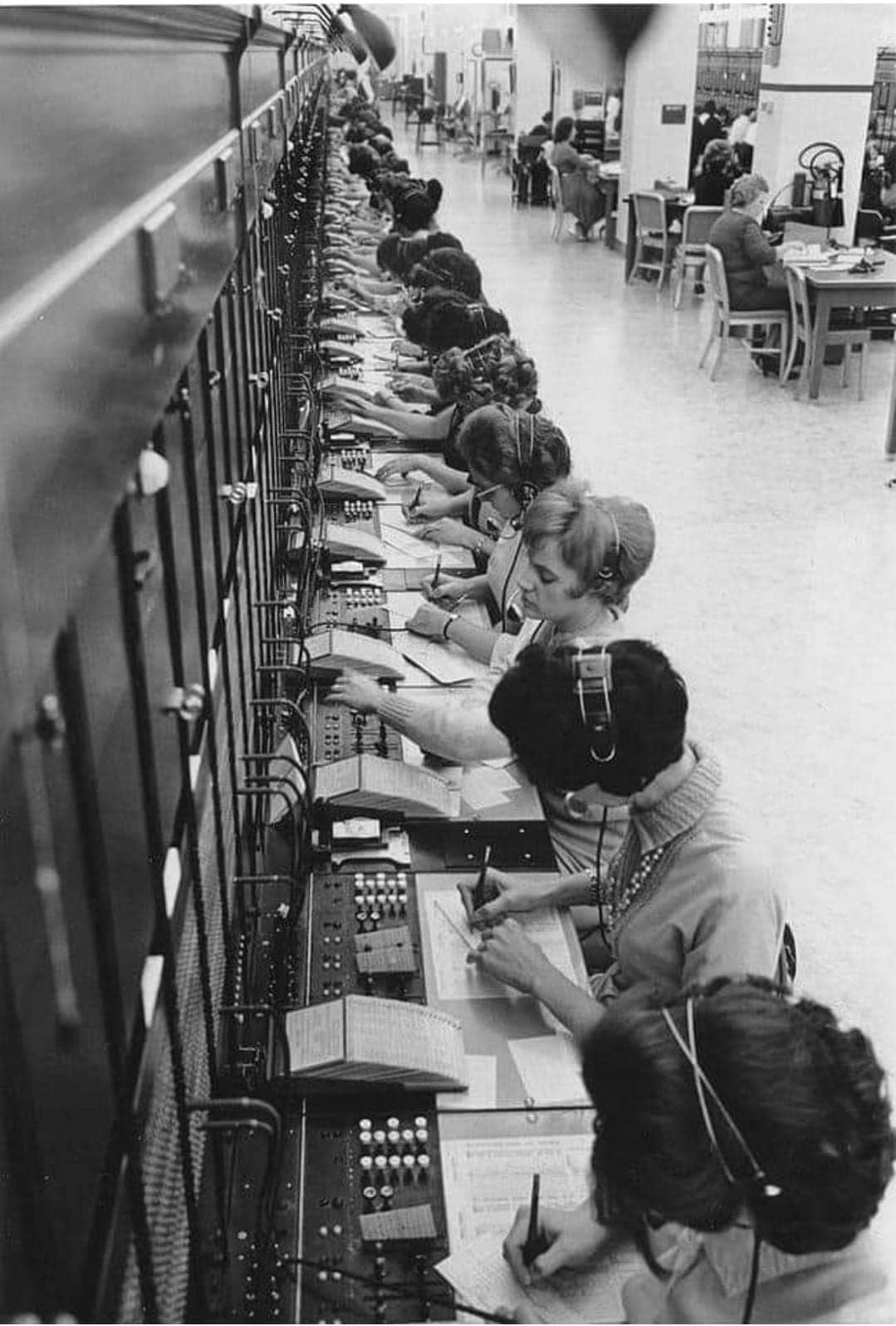 A row of women wearing headsets operate a large telephone switchboard, connecting calls and taking notes. The scene appears to be from a mid-20th-century office, with more workers visible in the background.