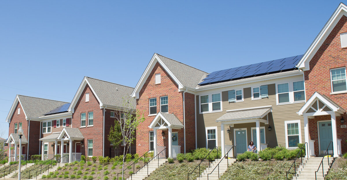 A row of modern, two-story townhouses with brick and siding exteriors, some with solar panels on the roofs. A person sits on the steps of one house, and neatly landscaped yards line the sidewalk.