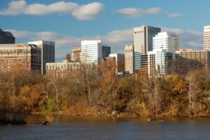 A river flows in the foreground with trees showing autumn foliage; behind them, a city skyline with modern office buildings rises under a blue sky with scattered clouds.