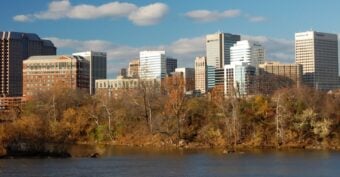 A river flows in the foreground with trees showing autumn foliage; behind them, a city skyline with modern office buildings rises under a blue sky with scattered clouds.