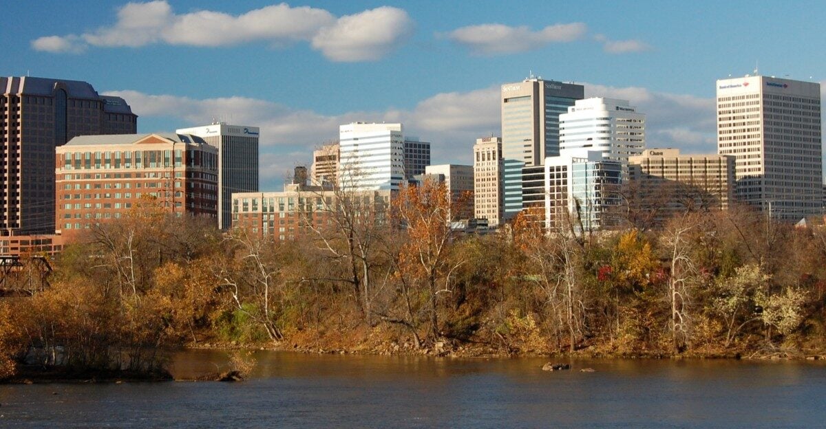 A river flows in the foreground with trees showing autumn foliage; behind them, a city skyline with modern office buildings rises under a blue sky with scattered clouds.