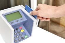 A person&rsquo;s hand inserts a blue credit card into a payment terminal with a keypad and display screen, suggesting a transaction or payment process.