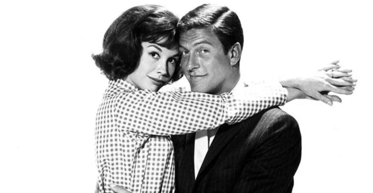 A woman in a checkered blouse hugs a man in a suit from the side. Both are smiling and looking at the camera against a plain white background.